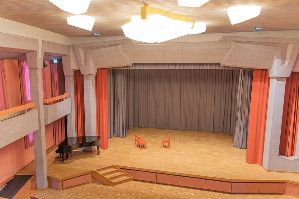 Das Bild zeigt eine Bühne auf der drei orange Stühle stehen. Die Bühnenvorhänge sind grau und orange. Auf der Bühne steht auch ein Piano.