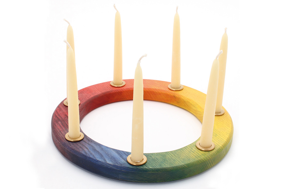 Das Bild zeigt einen bunten Holzring in dem sieben weisse Kerzen stecken.