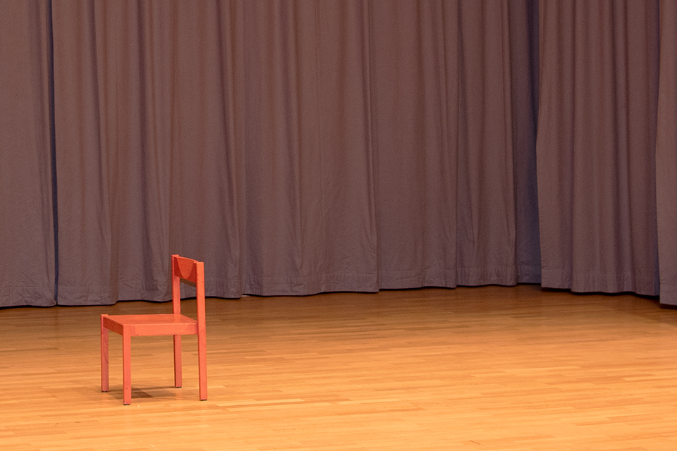 Das Bild zeigt einen organgen Stuhl auf einem Parkettboden und graue Bühnenvorhänge im Hintergrund.