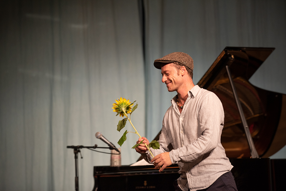 Das Bild zeigt einen lächelnden Mann mit einer Sonnenblume in der Hand. Im Hintergrund ist ein schwarzes Piano zu sehen.