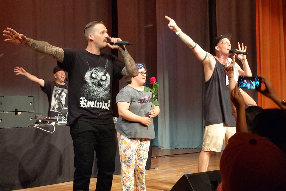 Das Bild zeigt zwei Sänger auf der Bühne in deren Mitte eine Frau mit einer Rose in der Hand steht. Im Hintergrund ist ein DJ zu sehen.