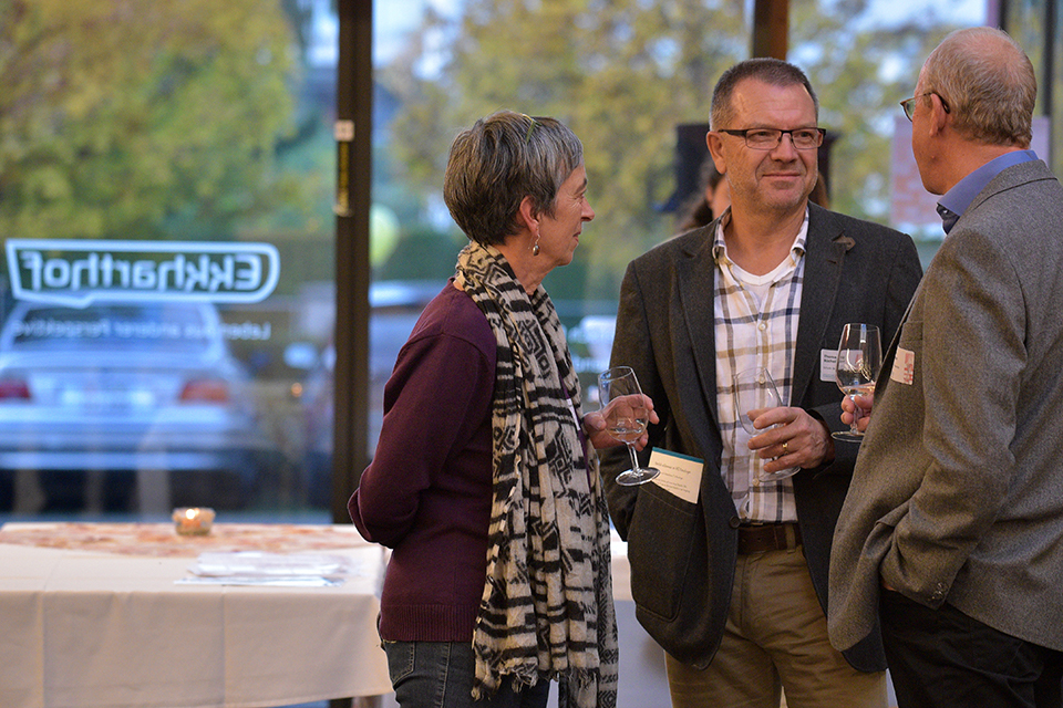 Das Bild zeigt zwei Männer und eine Frau miteinander im Gespräch. Alle Personen tragen ein Namensschild und halten leere Weingläser in den Händen.