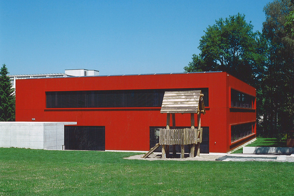 Das Bild zeigt ein rotes Gebäude mit Flachdach und grossen breiten Fenstern die mit schwarzen Storen geschützt sind. Vor dem Gebäude steht ein Holzspielhaus auf einer grossen Wiese.