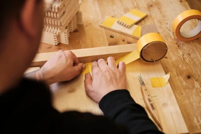 Man blickt über die Schulter eines Mannes, der mit einem Klebeband Holzstücke beklebt. Neben ihm liegt auf einem Brett ein Skalpell.