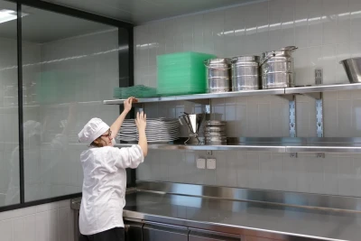 Man sieht eine Mitarbeiterin in weißer Schürze und Kochmütze vor einem Regal in der Großküche. Sie streckt sich nach Geschirr.