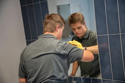 Man sieht einen jungen Mann von hinten, der gerade vor einem Spiegel in einem Bad steht und ihn mit einem Tuch putzt.