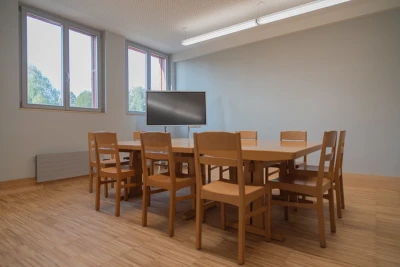Man blickt in einen Seminarraum. Darin steht ein Holztisch mit Holzstühlen und ein Monitor.