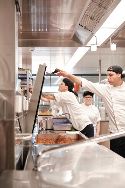 Drei Personen befinden sich in einer Gastronomie-Küche, sie tragen professionelle Kleidung und führen unterschiedliche Tätigkeiten aus.