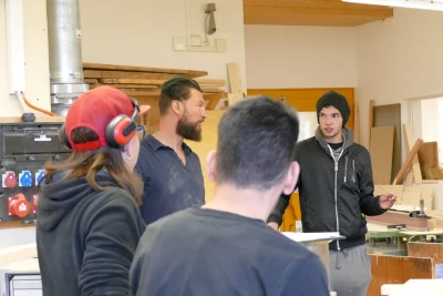 Zu sehen ist eine Gruppe junger Menschen, die in einer Werkstatt stehen und ein Gespräch führen. Eine trägt einen Gehörschutz.