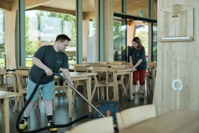Man blickt auf den Speisesaal, in ein Mensch mit einem Staubsauger über den Boden geht. Eine andere Person wischt Holztische ab.