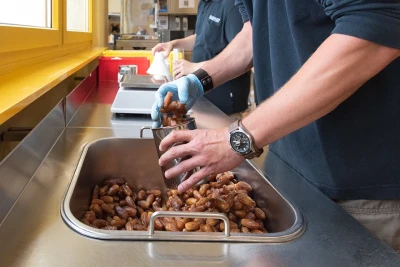 Man sieht die Unterarme und Hände von zwei Personen in einer Gastronomieküche, eine füllt Datteln in ein silbernes Gefäss ab.