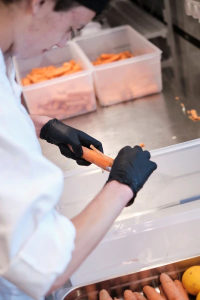Eine Person mit schwarzen Latexhandschuhen schält eine orange Karotte, man blickt ihr über die Schulter.