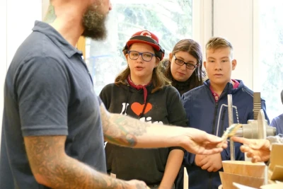 Zu sehen sind drei Jugendliche, eine davon trägt einen Gehörschutz. Ein Erwachsener zeigt etwas an einem Arbeitsgerät in der Holzwerkstatt.