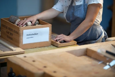 Zu sehen ist eine Frau mit Schürze, die in eine Holzkiste auf einer Werkbank greift. Sie ist beschriftet mit «Holzscheiben, ungeschliffen».