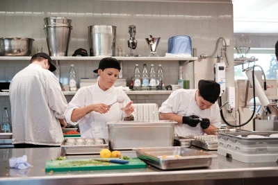 Drei Personen in einer Gastronomieküche führen unterschiedliche Tätigkeiten aus, die tragen professionelle Küchenkleidung.