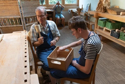 Zu sehen sind zwei Männer mit blauen Schürzen an einem Arbeitstisch in einer Werkstätte. Einer hat eine Holzkiste auf seinem Schoss.