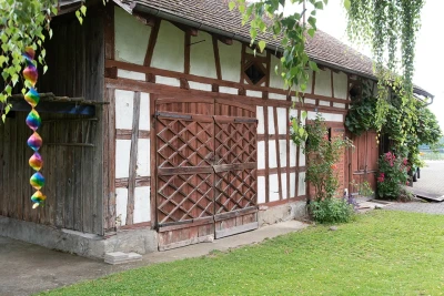 Zu sehen ist eine Fachwerk-Scheune. Sie hat drei grosse Holztore und auf der rechten Seite wachsen Pflanzen die Wand entlang.