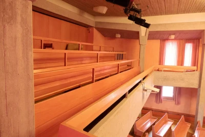 Man sieht die Tribühne mit festverbauten roten Sitzbänken. Darunter sind Holzbänke im Saal zu sehen.