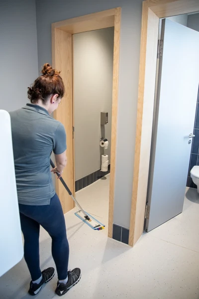 Zu sehen ist eine Frau mit einem Bodenwischer, die den Boden einer Toilette reinigt.