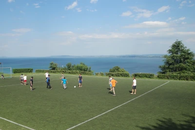 Man blickt über ein Fussbaldfeld, auf ihm spielen Personen Fussball, im Hintergrund blickt man über den Bodensee.
