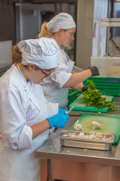 Zu sehen sind zwei Küchen­gehilfinnen, die an einer Arbeitsplatte stehen. Eine schält Knoblauch und eine schneidet Petersilie klein.