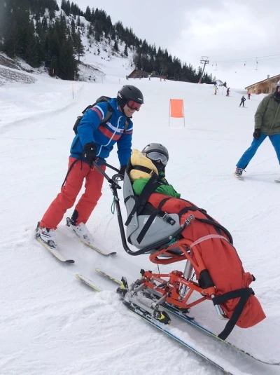 Zwei Personen fahren eine Skipiste hinab, einer auf Skiern, der andere sitzt in einem Bi-Ski und wird von der anderen person gehalten.