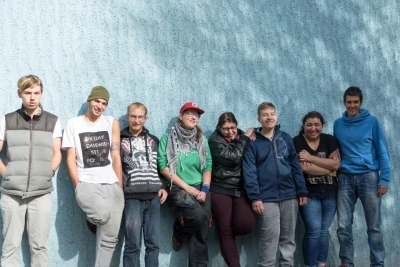 Zu sehen ist eine Gruppe von Jugendlichen, die sich vor einer blauen Wand in einer Reihe platziert haben und in die Kamera lachen.
