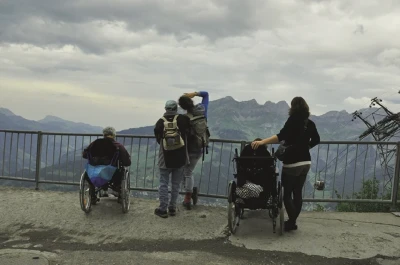 Fünf Persone, zwei davon im Rollstuhl blicken von einer Aussichtsplattform an einer Bergstation auf die Berge.