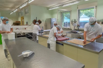 Man blickt in eine Bäckerei, mehrere Personen in professioneller Kleidung führen unterschiedliche Tätigkeiten aus.