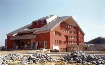 Man blickt auf ein rotes Hügelhaus mit auskragenden Dächern. Vor dem Haus liegen einige Haufen an Bausteine.