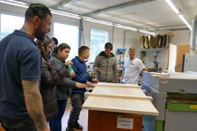 Man blickt auf eine Holzplatte in einer Holzwerkstatt um die eine Gruppe junger Menschen stehen. Ein Mann gestikuliert.