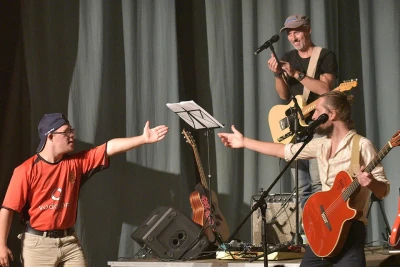 Man sieht eine Band mit zwei Gitarrenspielern, Lautsprechern und Mikrofonen, eine dritte Person deutet auf einen Gitarrenspieler.