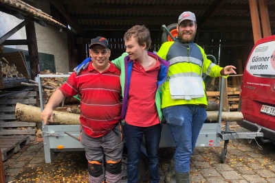 Zu sehen sind drei Männer in Arbeitskleidung, die sich im Arm halten und vor einem Anhänger mit Holzstämmen stehen.