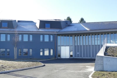 Zu sehen ist das Gebäude der Turnhalle mit bläulicher Fassade. Der Blick ist hin zum Eingang gerichtet.