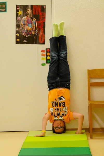 Ein Kind mit Brille macht einen Kopfstand auf einer grünen Matte. Auf die Wand dahinter sind Poster und Zettel geklebt.