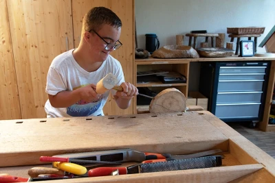 Zu sehen ist ein Junge mit einer eingespannten Holzscheibe an einer Werkbank. Er schlägt mit einem Holzhammer auf ein Stemmeisen.