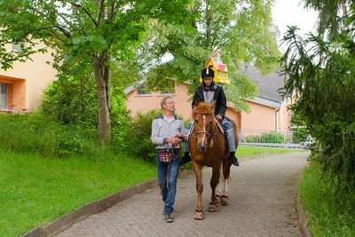 Auf einem Weg reitet ein Kind auf einem Pferd dem Betrachter entgegen, es wird von einer erwachsenen Person geführt.