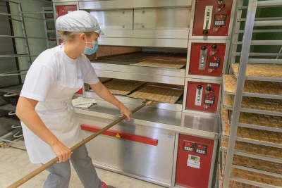 Eine Person die in einer Bäckerei arbeitet schiebt ein Blech Kekse in einen Offen, sie trägt professionelle Kleidung.