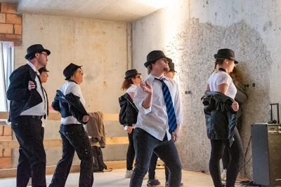 Man sieht die Tanzgruppe beim Tanzen, alle tragen Hüte, Anzüge und Krawatten, sie ziehen gerade ihre Jackets aus.