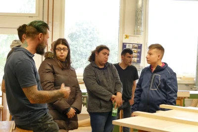 Man blickt auf einen Mann, der gerade zu einer Gruppe von Jugendlichen in der Holzwerkstatt spricht. Im Hintergrund sind Werkbänke zu sehen.